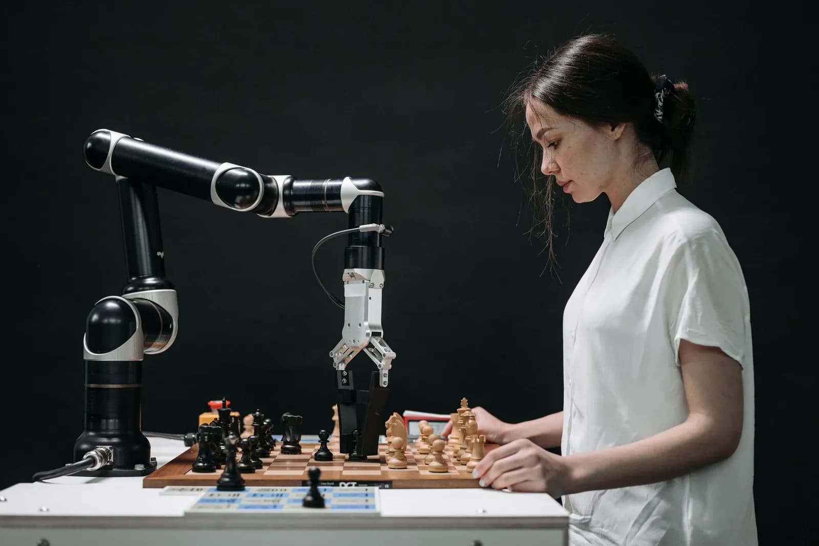 A young woman in a white blouse playing chess against a robot arm.