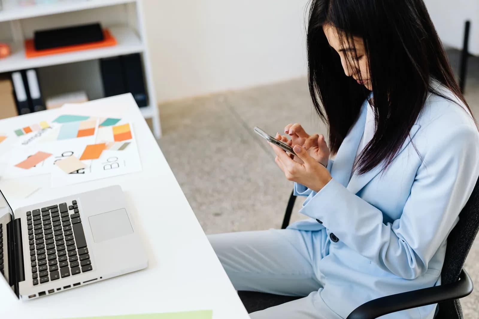 Businesswoman using smartphone at desk with laptop and coffee cup.