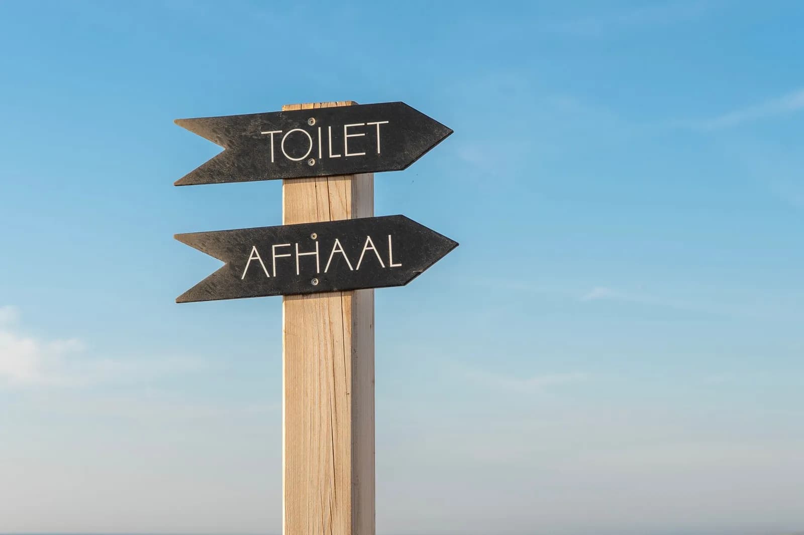 Directional signpost with 'Toilet' and 'Afhaal' against a clear blue sky background.