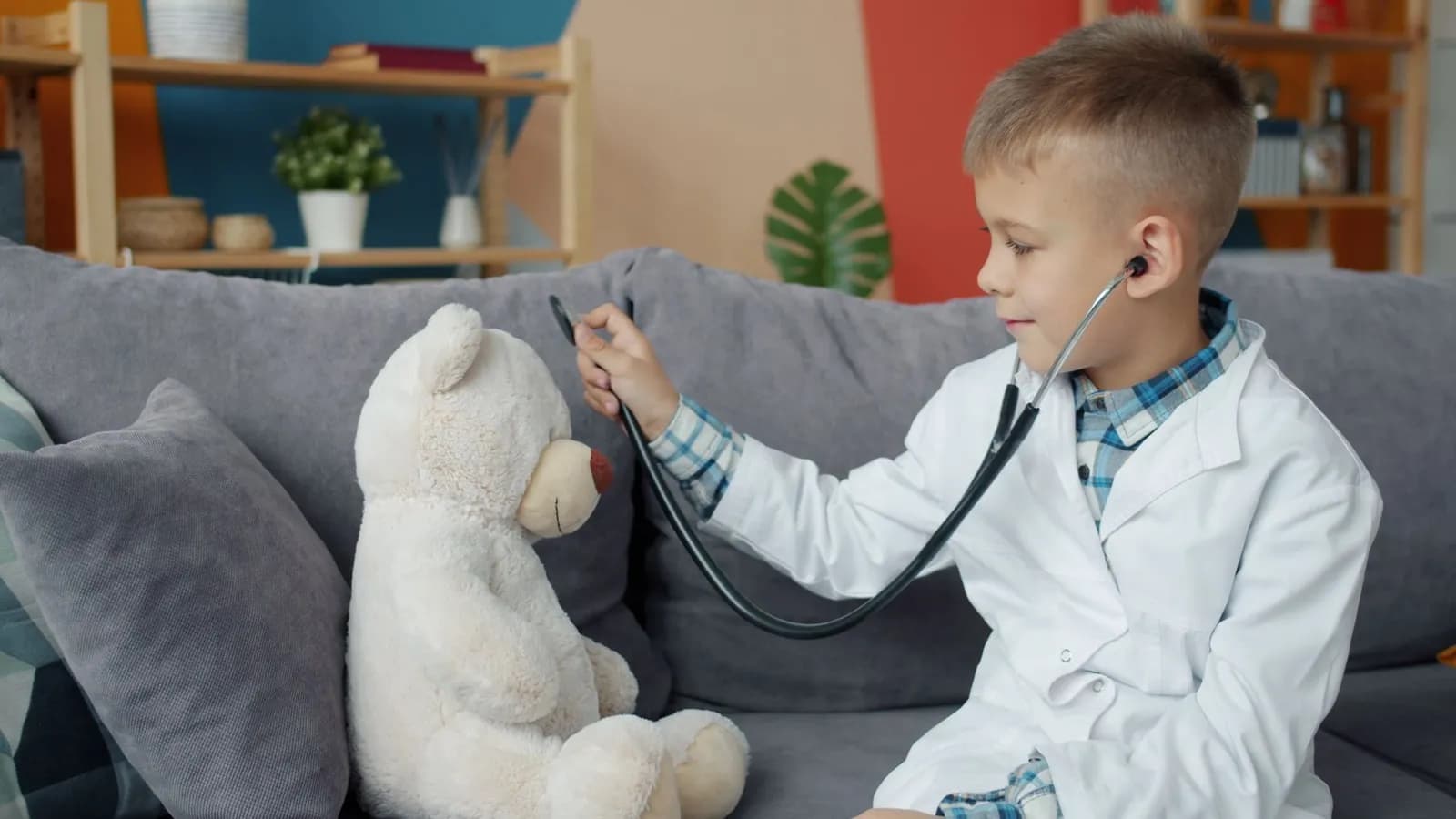 A boy plays doctor with a teddy bear, using a stethoscope on the couch indoors.