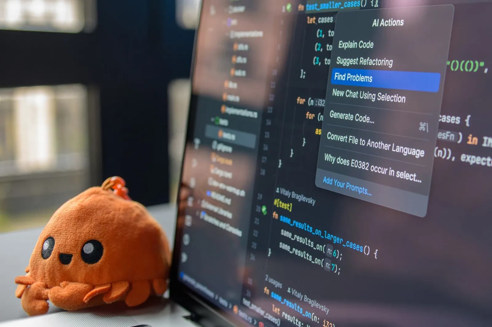 A laptop screen showing a code editor with a cute orange crab plush toy beside it.