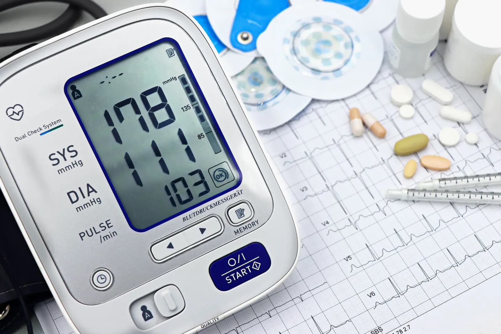 Close-up of a digital blood pressure monitor with ECG printout, pills, and medical tools.