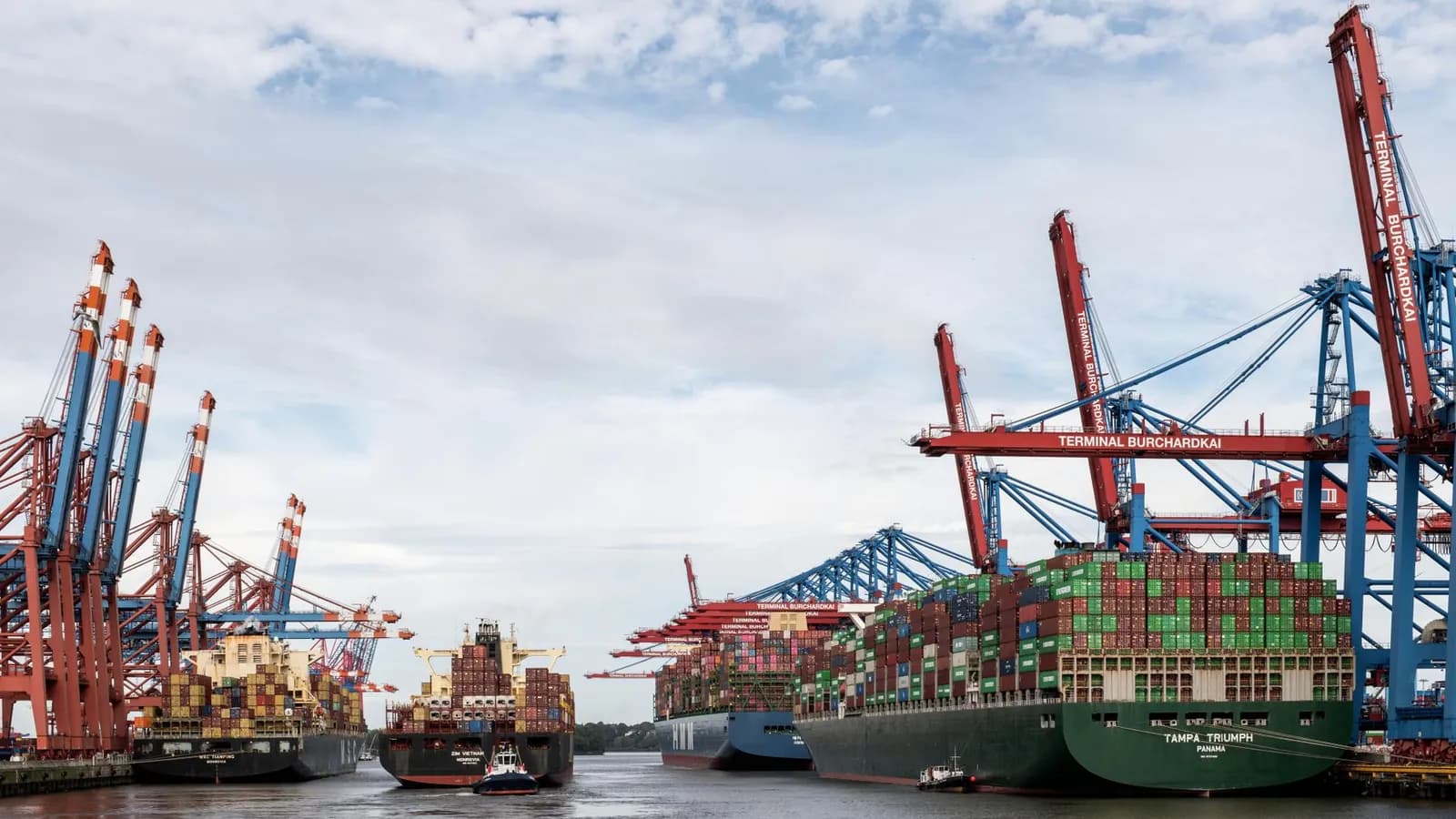 Container ship docked at port under cranes during twilight, illuminated against the evening sky.