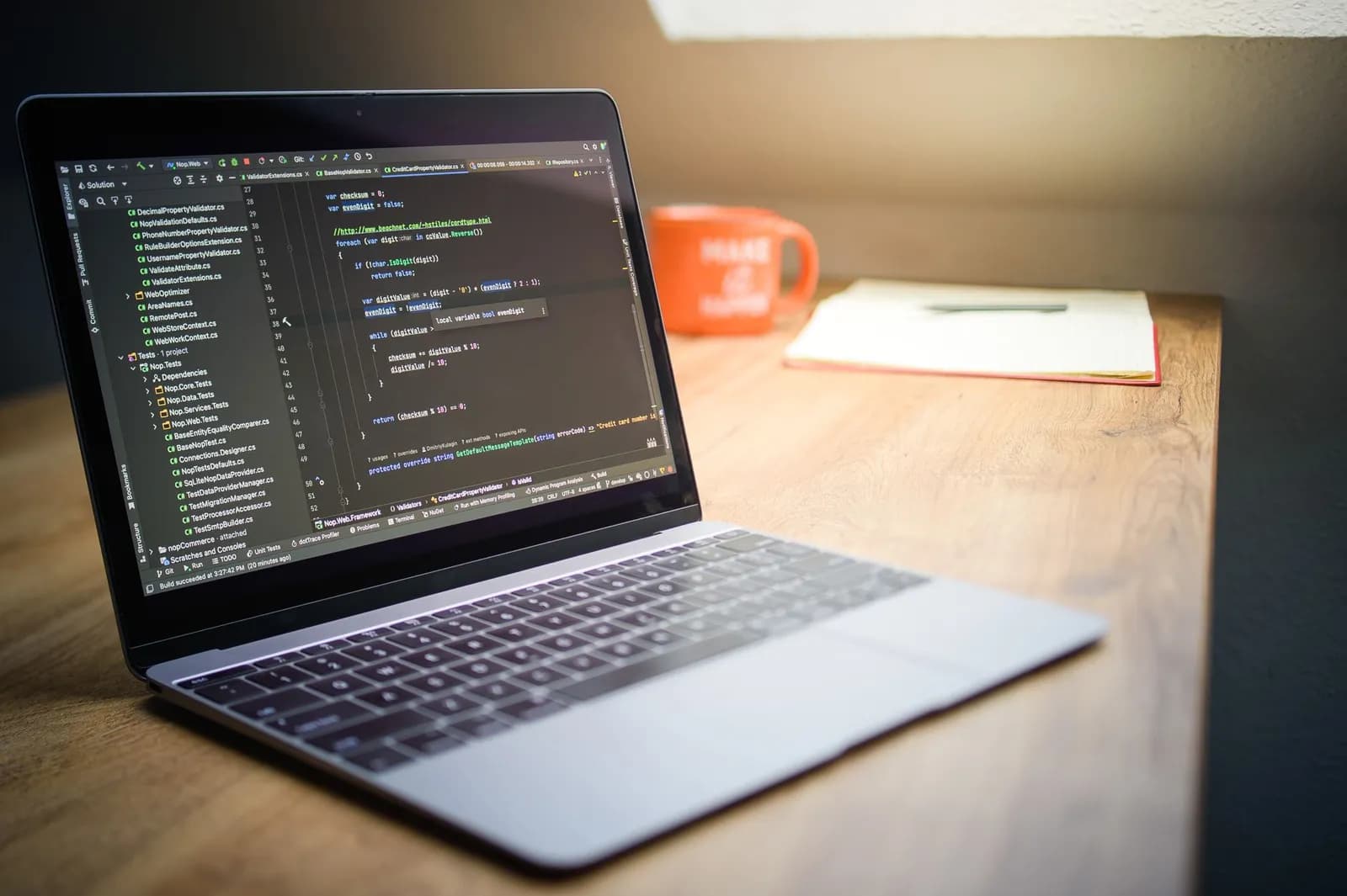 Laptop showcasing code on a desk with a notebook and mug, emphasizing a modern workspace setting.