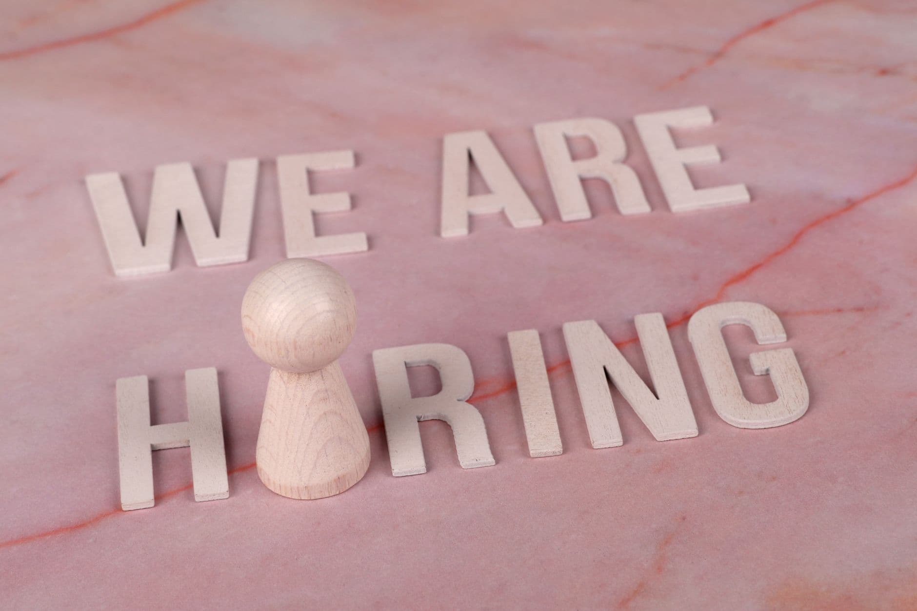 Wooden letters spelling 'We Are Hiring' on a pink marble surface with a pawn piece.