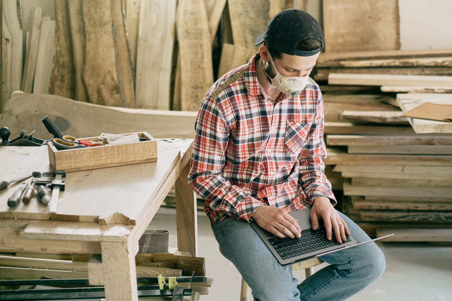 A carpenter in a workshop using a laptop, surrounded by tools and wood.