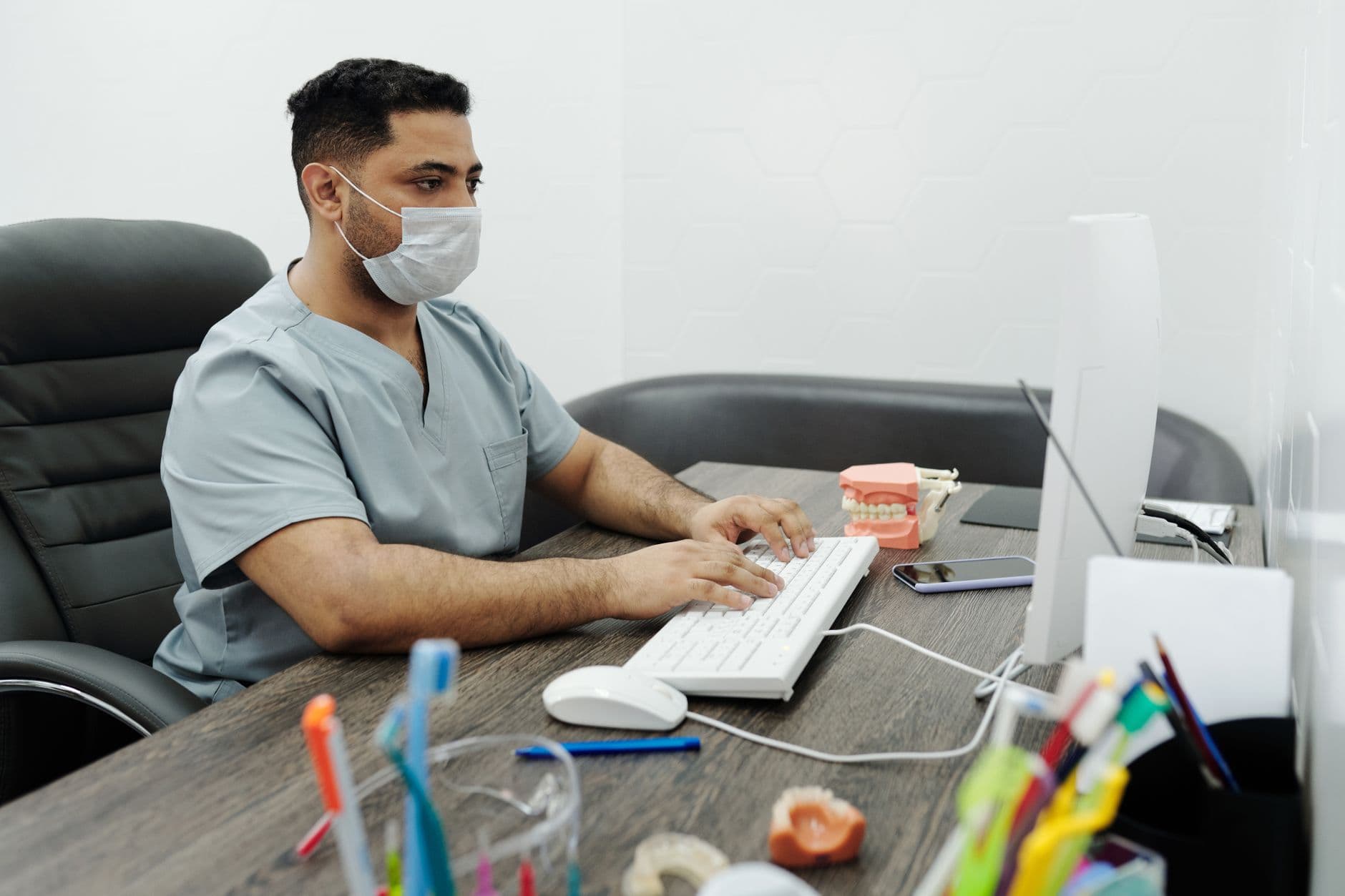 Dentist wearing mask working on computer at office desk with dental model.