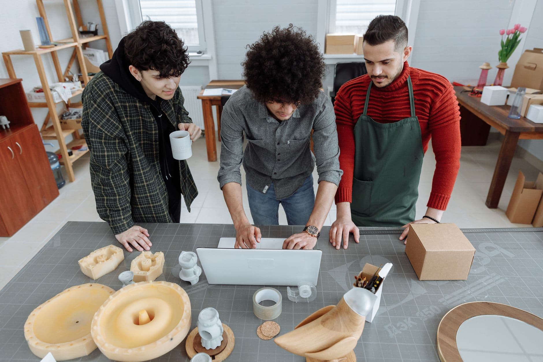Three men in a workshop setting working together with a laptop and various crafting materials on the table.