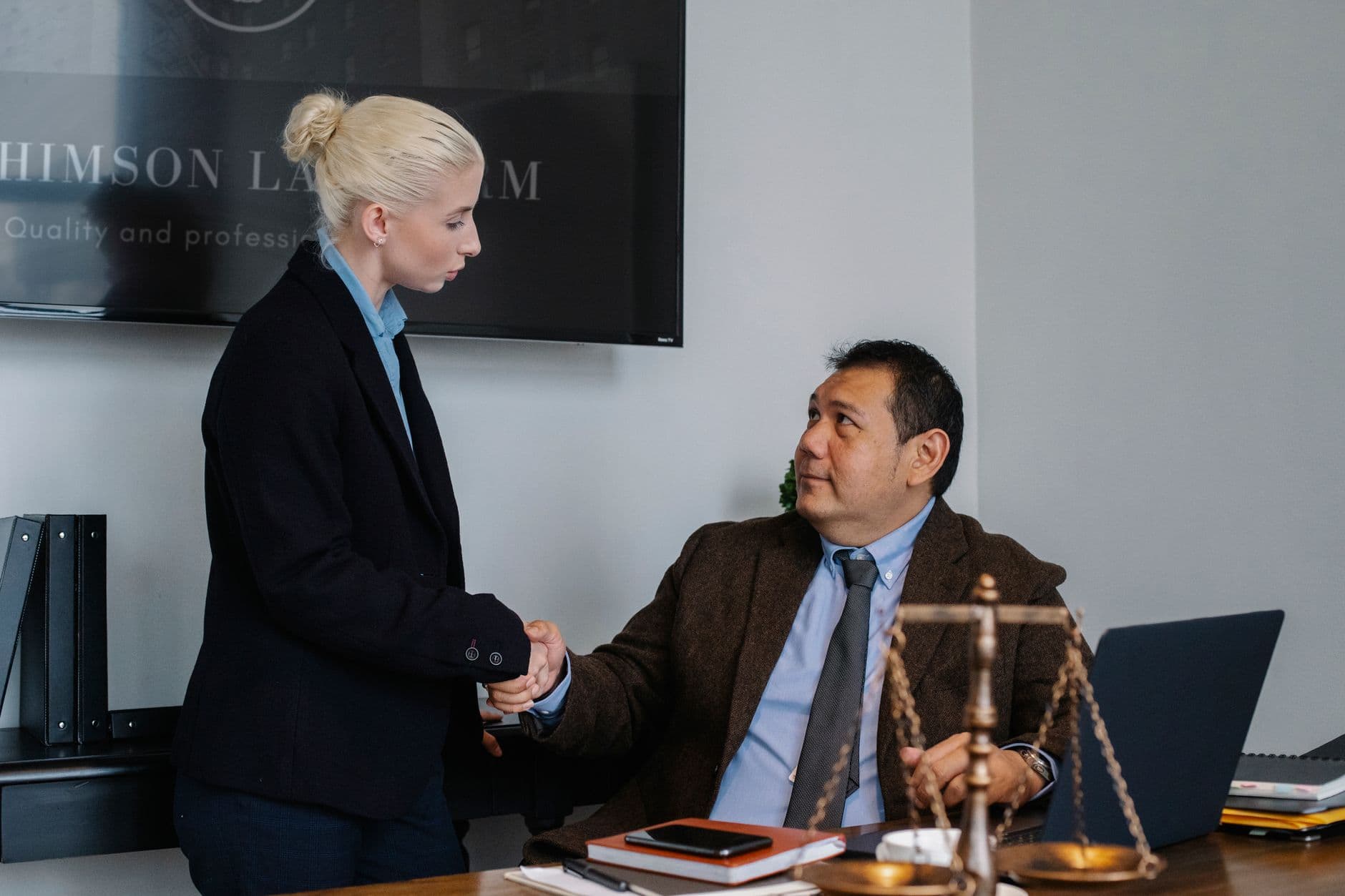 Multiracial coworkers in formal outfit shaking hands near table with scales of justice