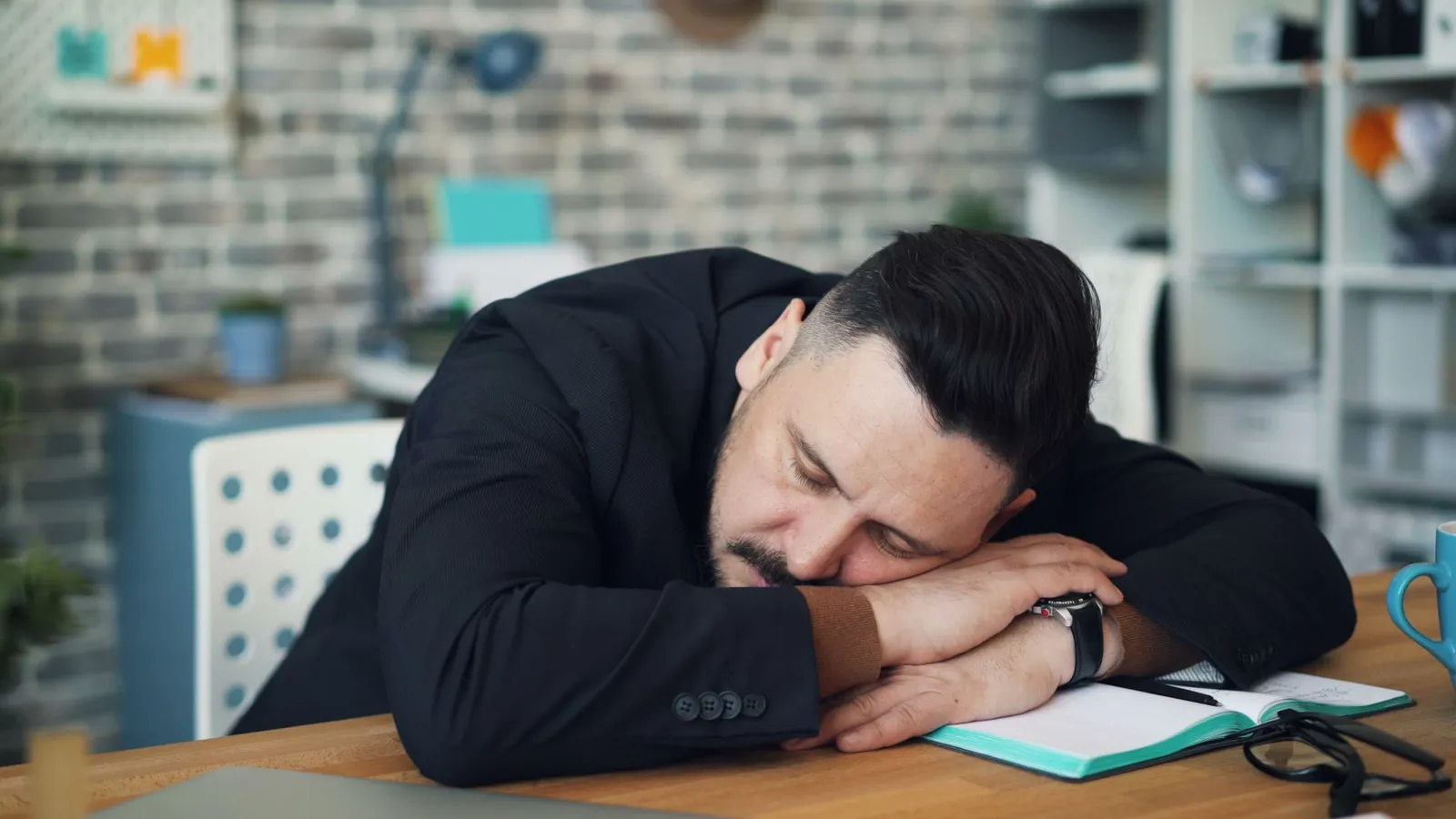 Exhausted man in office resting on desk with notebook and coffee mug.