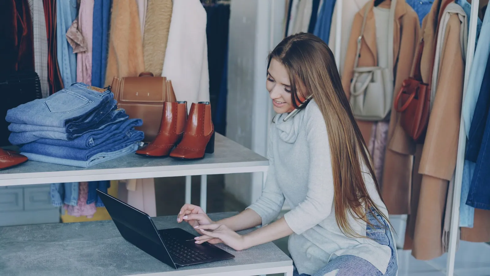 Woman in boutique talks on phone with laptop, surrounded by clothes.
