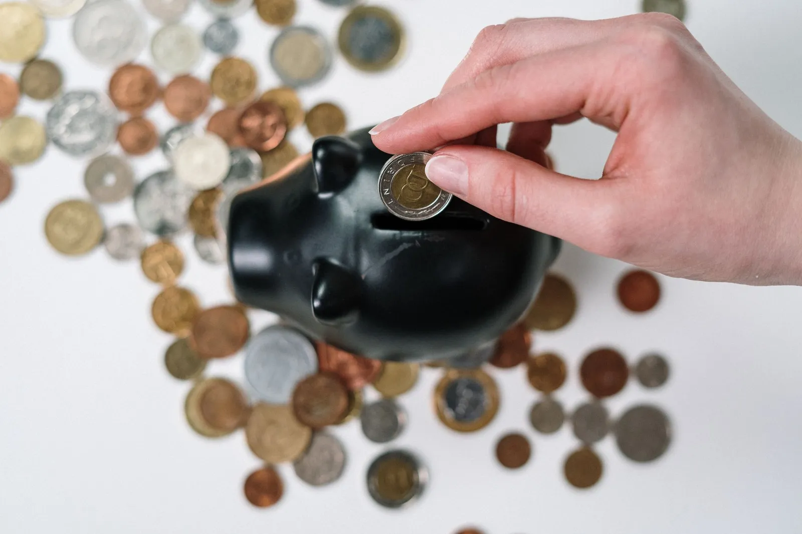 Close-up of a hand inserting a coin into a black piggy bank with scattered coins on a white background.