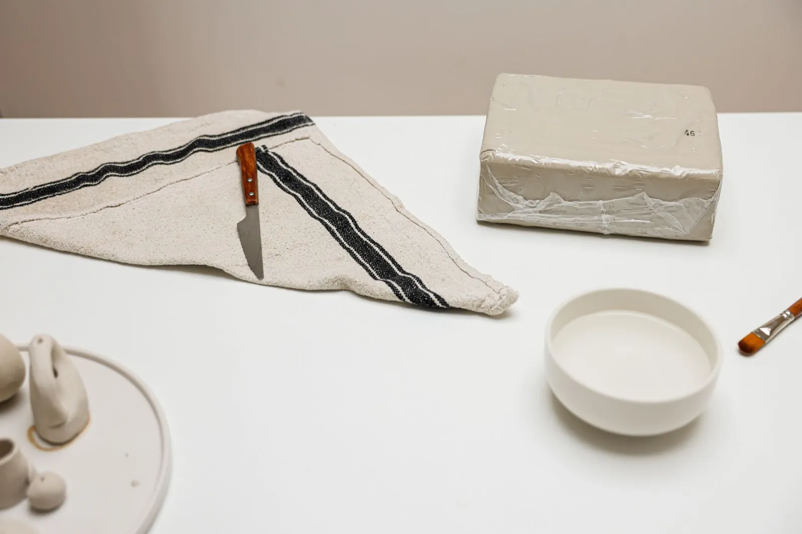 A minimalist ceramic artist's workspace featuring tools, a clay block, a bowl, and figurines on a white surface.