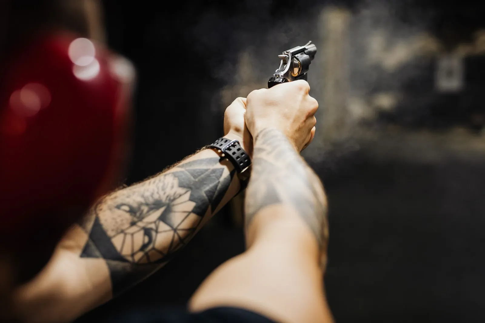 Close-up of tattooed arms aiming and firing a revolver in an indoor shooting range.