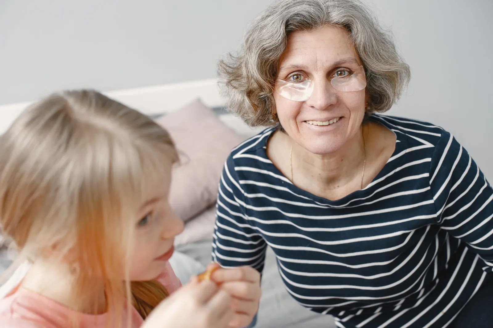 Smiling elderly woman with a child indoors, wearing under-eye patches in a cozy setting.
