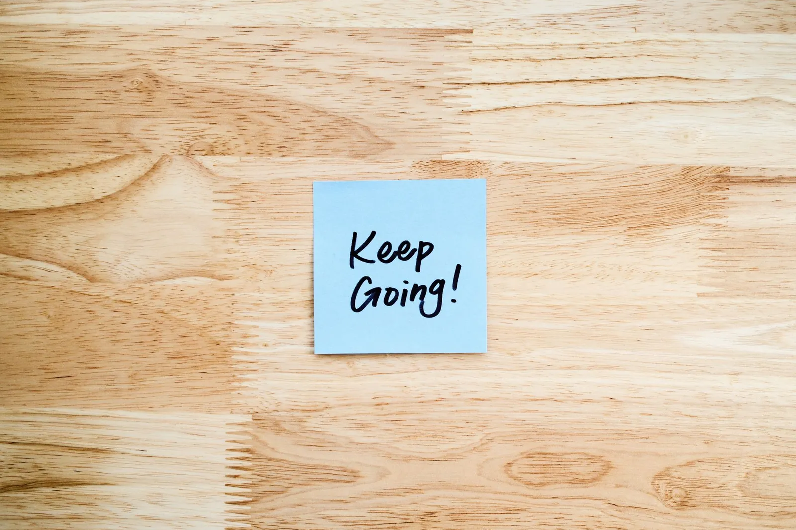 A blue sticky note with 'Keep Going!' message on wood background.